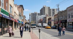 A street-level view of Main Street in Fort Lee, New Jersey, a bustling commercial area. On the left, a row of storefronts with signs for 'K-Food Market', 'Main Street Cafe', and 'Fort Lee Pharmacy' are visible, with pedestrians walking by. The road features moderate traffic, including cars, a bus, and delivery trucks. In the background, tall apartment high-rises dominate the skyline under a clear blue sky with a few clouds. Other storefronts like 'Banco Popular' and 'CityMD' are on the right side of the street.