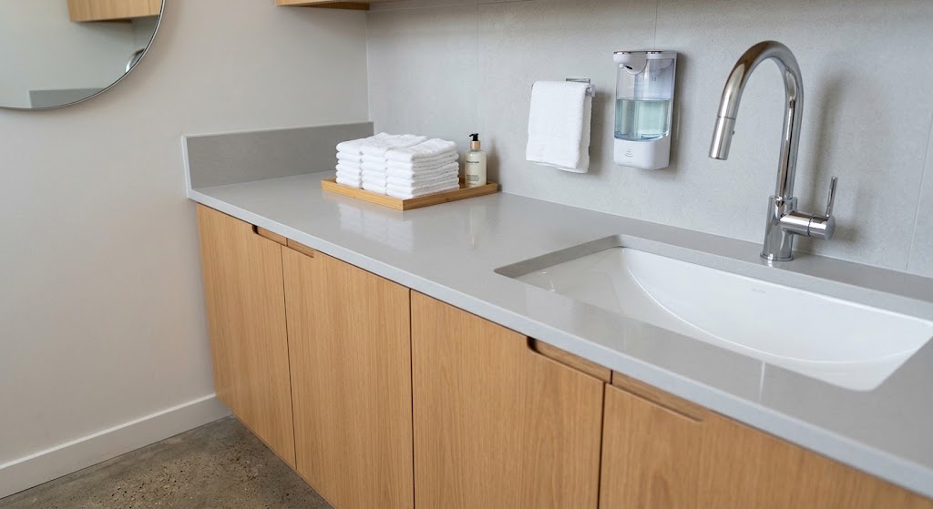 Modern commercial bathroom vanity featuring light oak wood cabinets and a sleek grey quartz countertop with an integrated white sink. The setup is accessorized with a modern chrome faucet, a wall-mounted automatic soap dispenser, and a wooden tray holding fresh white towels and a premium hand wash bottle.